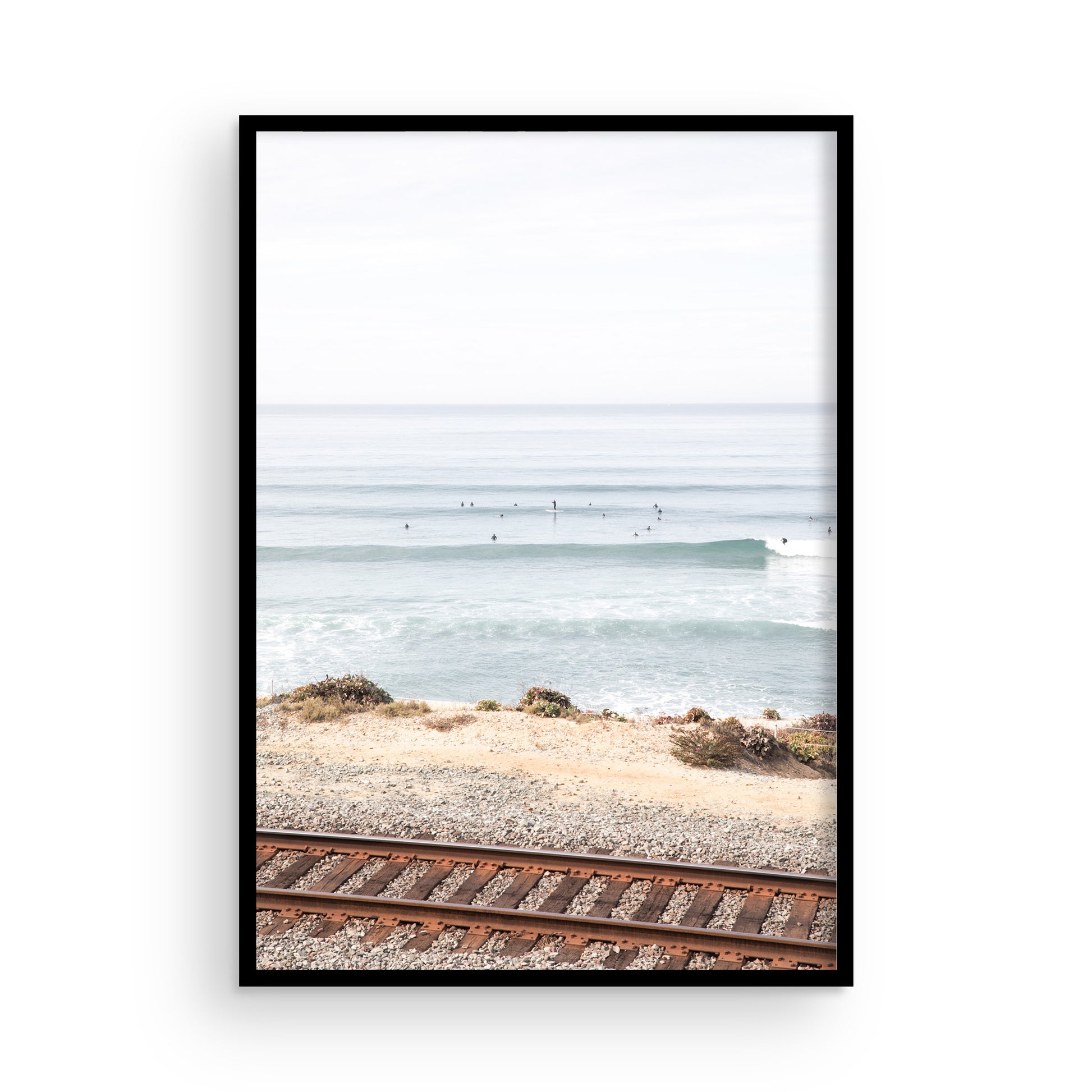A framed print of a beach scene with waves and surfers, displayed on a train track in Del Mar, San Diego. Photograph in minimalist mock up.