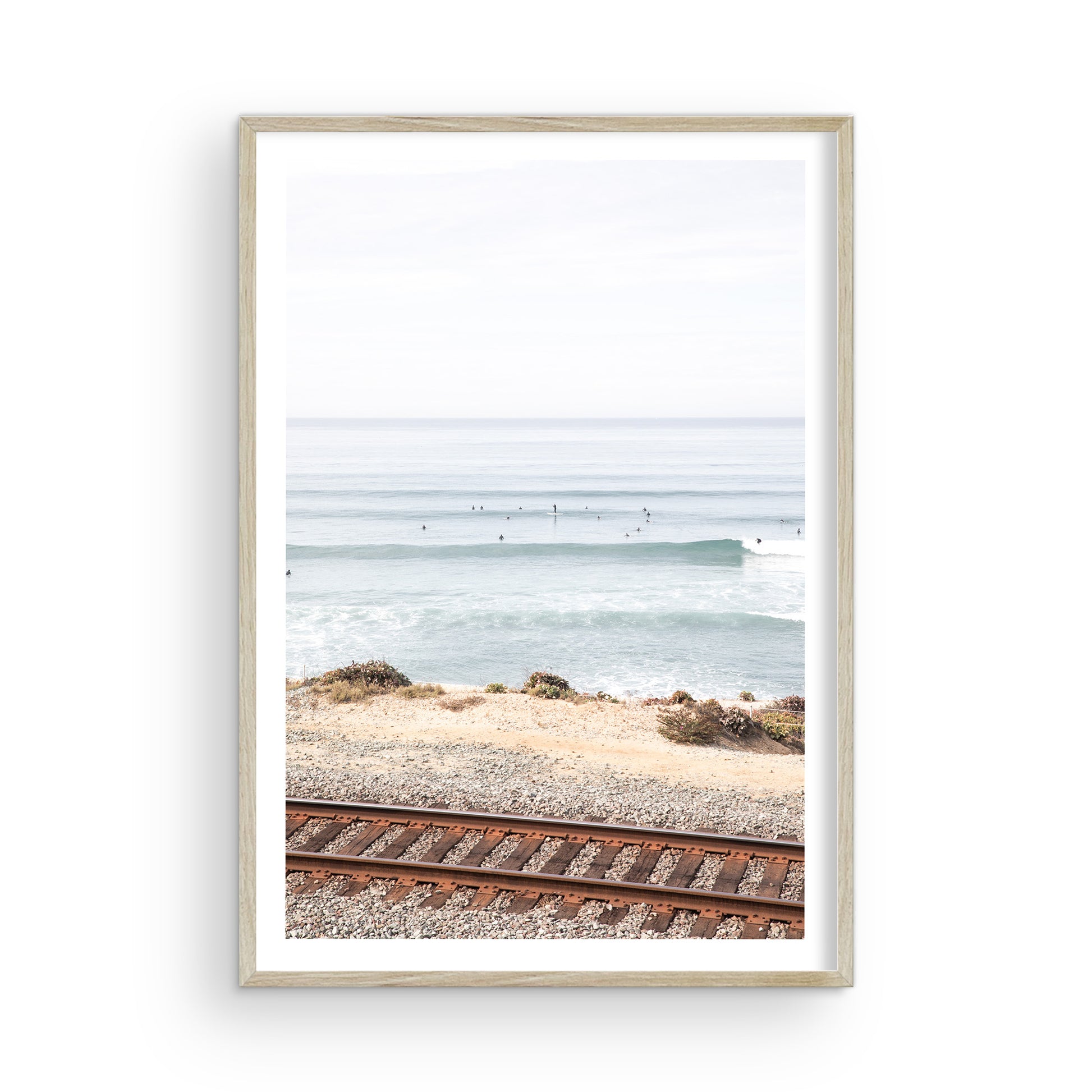 A framed print of a beach scene with waves and surfers, displayed on a train track in Del Mar, San Diego. Photograph in minimalist mock up.