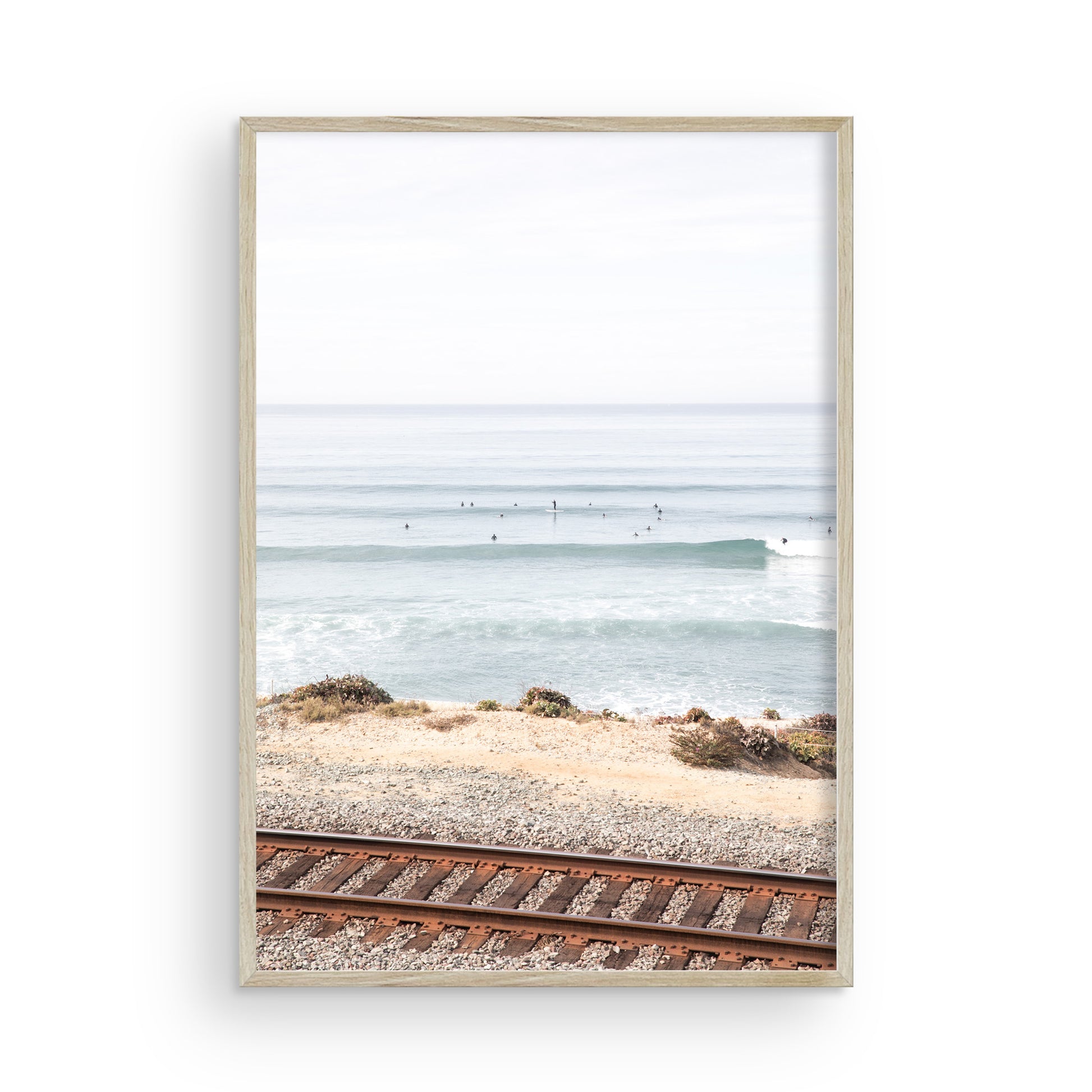 A framed print of a beach scene with waves and surfers, displayed on a train track in Del Mar, San Diego. Photograph in minimalist mock up.