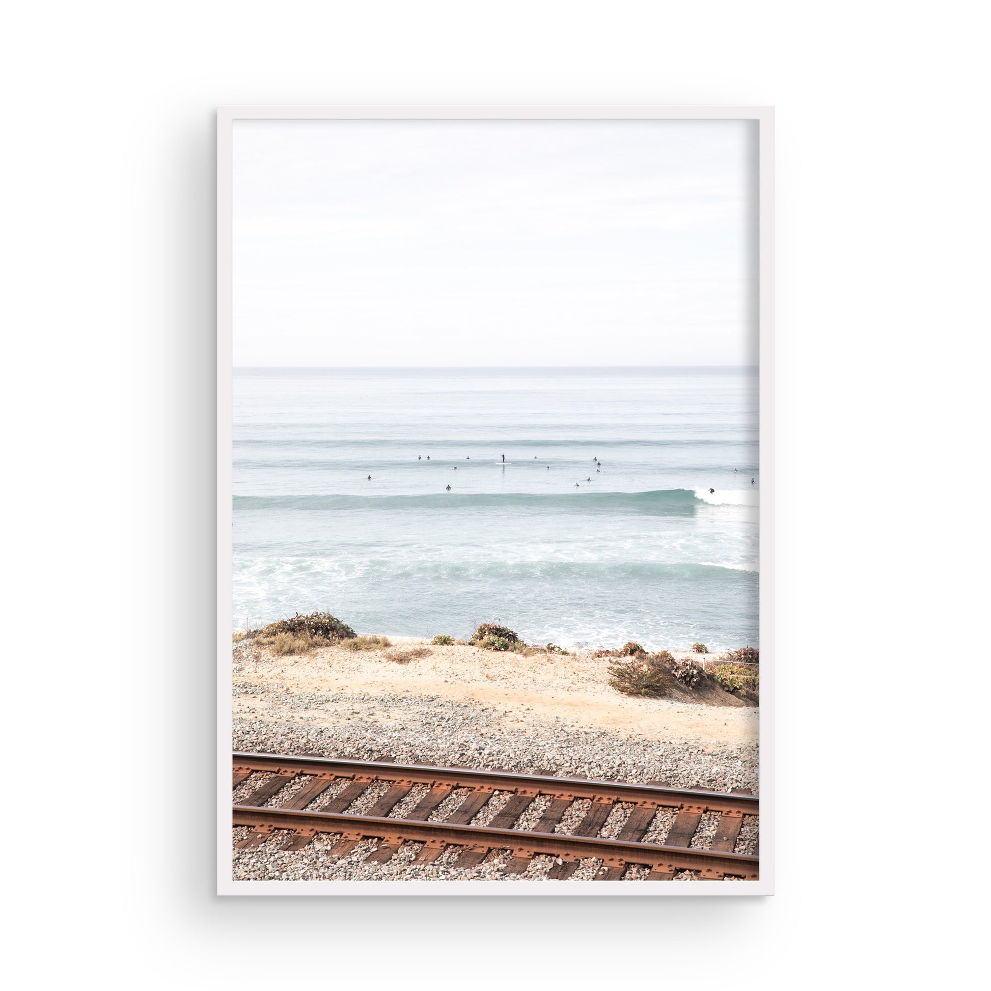 A framed print of a beach scene with waves and surfers, displayed on a train track in Del Mar, San Diego. Photograph in minimalist mock up.