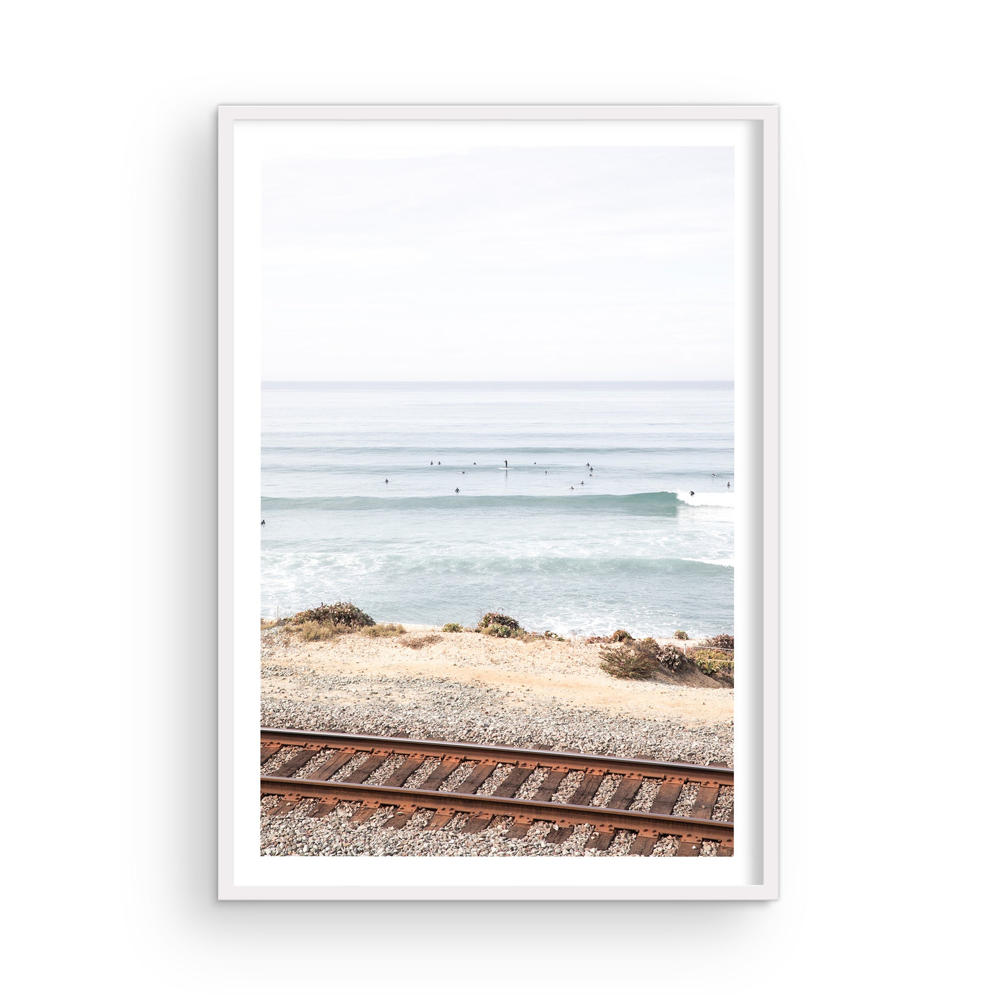 A framed print of a beach scene with waves and surfers, displayed on a train track in Del Mar, San Diego. Photograph in minimalist mock up.