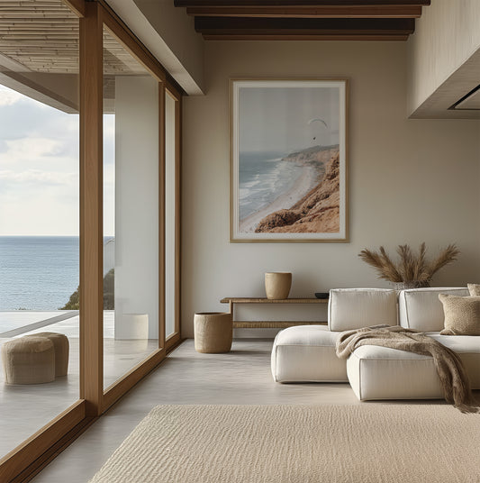 Fine art of Torrey Pines, San Diego California vertical print of hazy high-angle landscape beach photography captures a light blue ocean meeting a sandy beach with many beach goers. To the right, tall tan and brown cliffs rise as iconic coastal scenery. A white paraglider with a pilot flies in the upper right against a pale pastel sky, a travel photography moment. The scene emphasizes the cliffs, beach, and the paraglider's presence in the expansive large beach art view. Photograph in minimalist mock up