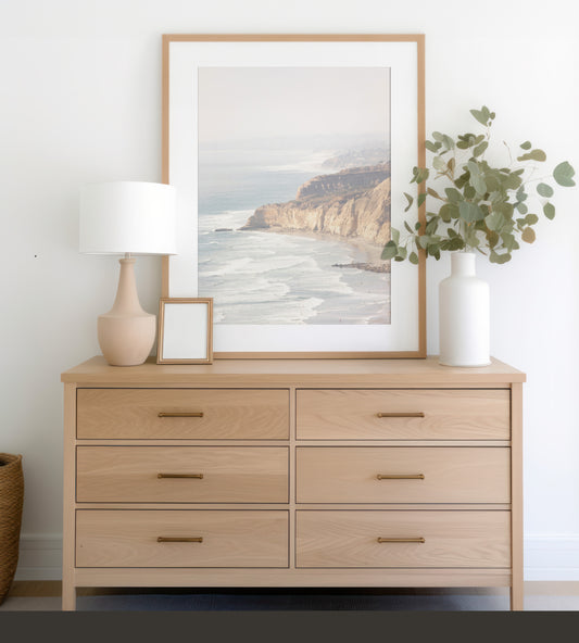 Fine art of Torrey Pines, San Diego California vertical print of hazy high-angle landscape beach photography captures a light blue ocean meeting a sandy beach with many beach goers. To the right, tall tan and brown cliffs rise as iconic coastal scenery. Photograph in minimalist framed mock up.