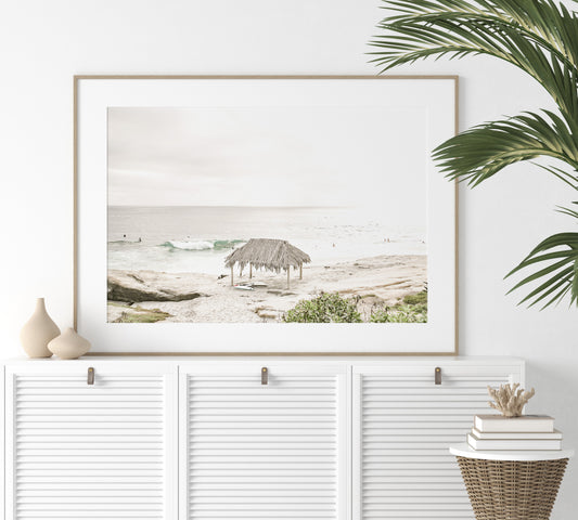 Fine art of WindanSea Beach, La Jolla California horizontal landscape print by Southern California photographer Kim Dybczak of Salt Creek Prints, featuring a bright, slightly desaturated image shows Windansea Beach in La Jolla, California. A thatched beach hut stands on the sandy shore in the left foreground. Further down the beach, a surfer carrying a teal surfboard walks towards the ocean where several other surfers are visible in the water. Photograph in minimalist framed mock up