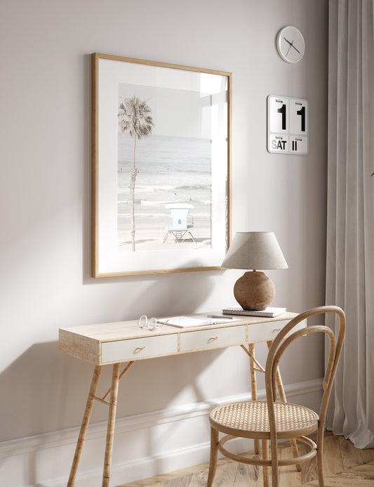 Dreamy coastal palm tree print featuring a tall palm tree with shaggy trunks in the foreground and a pastel blue lifeguard tower centered in the distance. This vertical tropical print is captured at Oceanside Beach on the Southern California coast, is bathed in bright, hazy sunlight, creating a pastel beach art effect. Photograph in minimalist mock up.