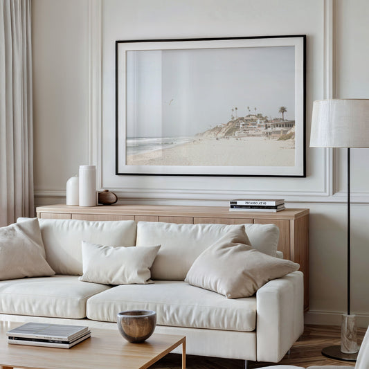 A bright, minimalist photograph of Moonlight State Beach in Encinitas, California, showing a wide sandy beach, gentle ocean waves, and residential buildings atop a cliff on the right, with a seagull flying in the hazy sky. Photograph in minimalist mock up