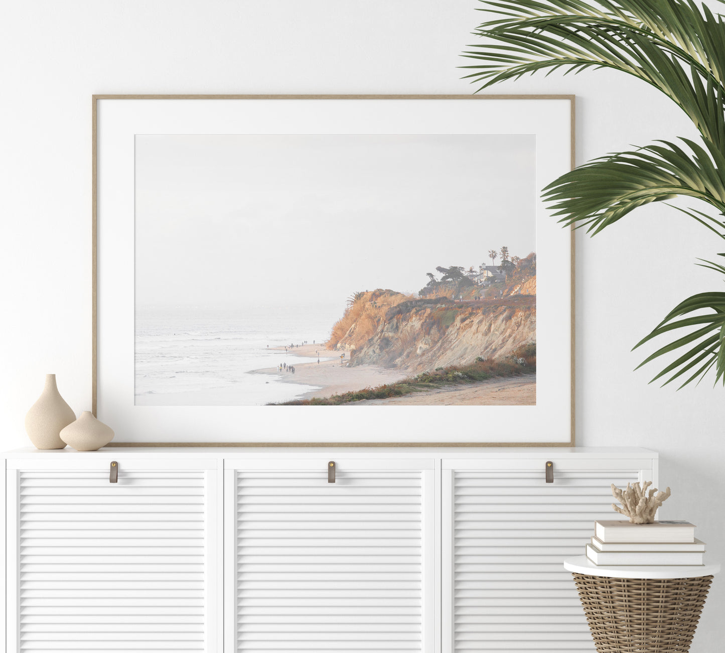 Fine art of Torrey Pines, San Diego California horizontal print of hazy high-angle landscape beach photography captures a light blue ocean meeting a sandy beach with many beach goers. To the right, tall tan and brown cliffs rise as iconic coastal scenery. Photograph in minimalist framed mock up.