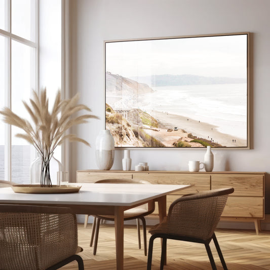A slightly hazy, high-angle Landscape Beach Print captures a wide beachscape print where a light blue ocean waves meet a sandy shore dotted with numerous people enjoying the Summer Vibes. Gentle white waves roll towards the beach. Blue Lifeguard Towers line the coast in the far distance, of this framed landscape beach print. The overall lighting suggests a bright, overcast travel photography day on the San Diego coast. Photograph in minimalist mock up