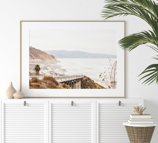 Captivating San Diego coast scene, presented as horizontal beach art. This piece of travel photography features a prominent bridge extending over a wide sandy beach with calm ocean waves. A coastal road winds along the base of rugged cliffs, with a lone coastal palm tree adding a touch of tropical essence to this Southern California seascape print. Photograph in minimalist mock up
