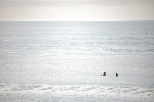 Horizontal minimalist ocean art with a close-up blue waves print of water, featuring subtle ripples and soft textures, ideal as a neutral ocean print, two surfers sit and watch the waves.