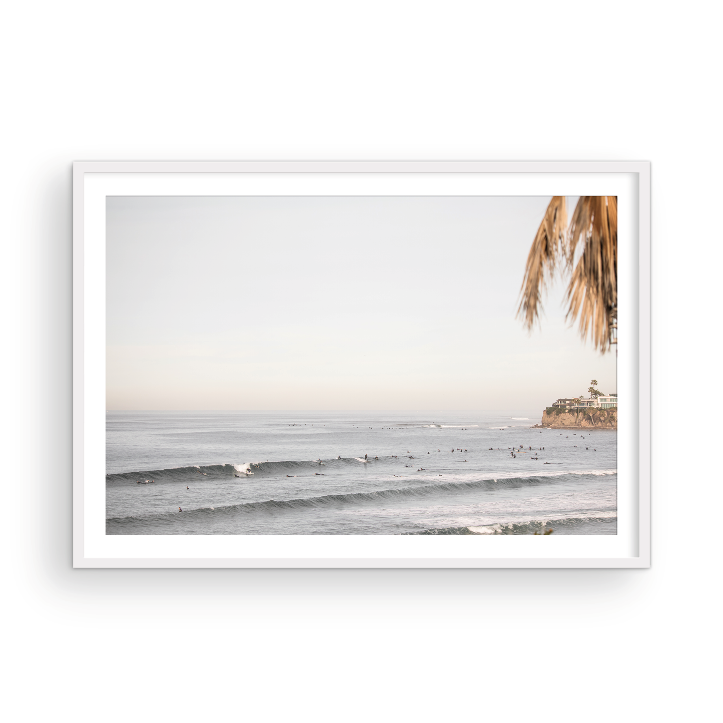 Landscape beach photography of Tourmaline Beach, Pacific Beach, California, with waves and many surfers, framed by a palm tree in the corner. This ocean print captures casual surf vibes. Photograph in minimalist mock up.