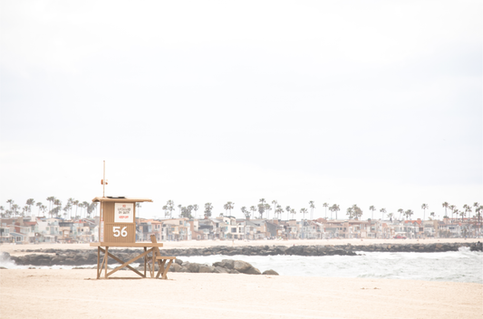 Landscape beach photography of Newport Beach, featuring lifeguard tower 56 on the sand with ocean waves and coastal houses in the background. A great Newport Beach photography print.