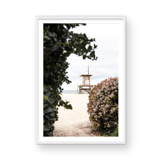 Vertical coastal photography of a Newport Beach lifeguard tower on the sand, framed by lush green and flowering foliage. Photograph in minimalist mock up.