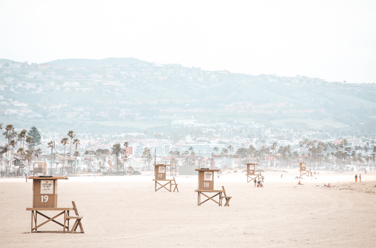 Landscape beach photography of Santa Monica State Beach with multiple lifeguard towers on the vast sandy expanse and hazy hills in the background.