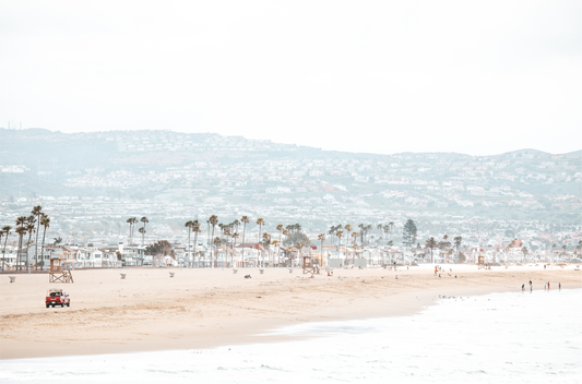 A wide landscape coastal photography shot of Newport Beach, California, featuring a red lifeguard truck, sandy beach, palm trees, and hazy mountains in the distance.