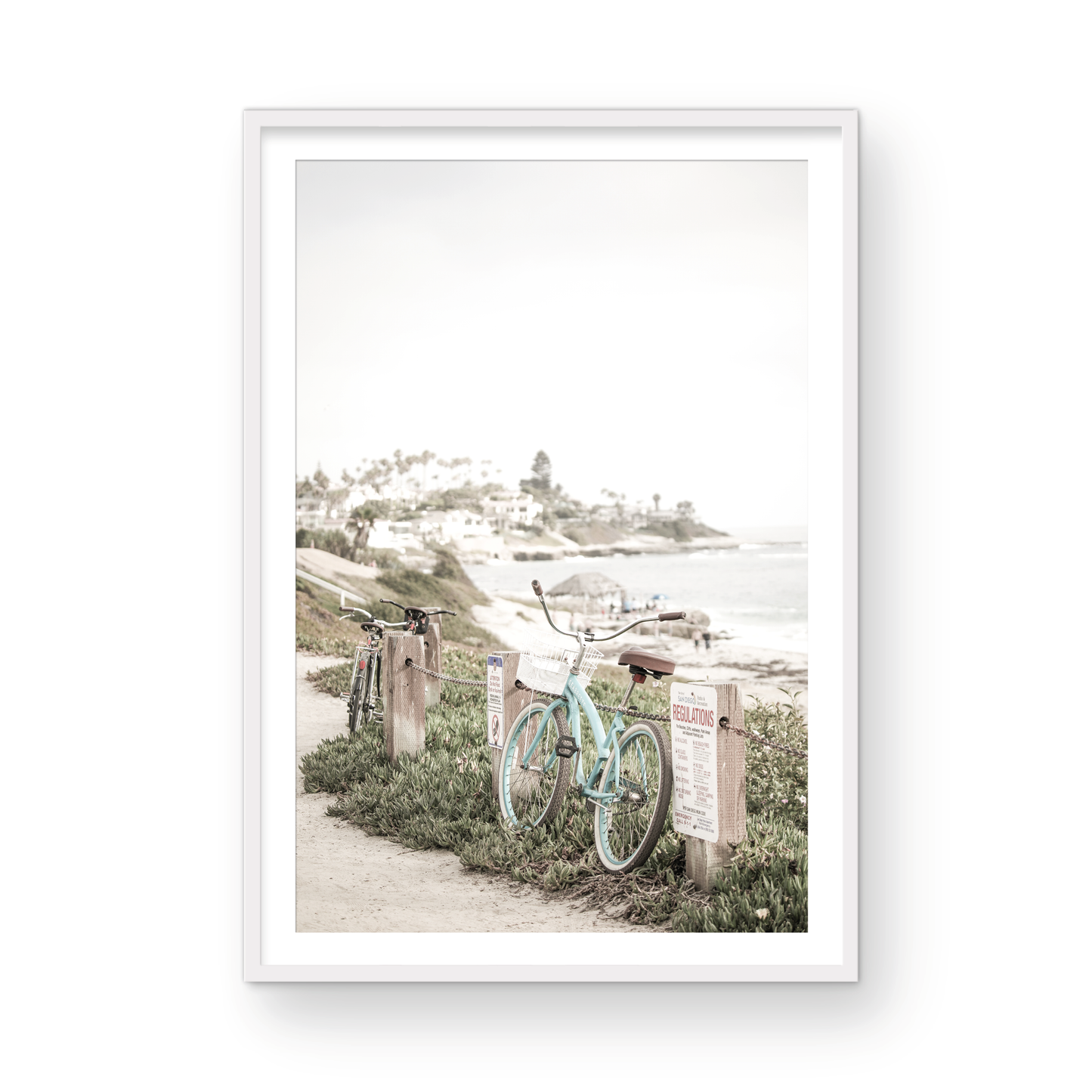 Fine art of horizontal landscape print by Southern California photographer Kim Dybczak of Salt Creek Prints, the image shows a light blue pastel beach cruiser leaning against a wooden post and chain fence. Another bicycle is visible behind it. The beach is in the background with people and a beach hut, located in Windansea Beach in La Jolla, California. Photograph in framed minimalist mock up