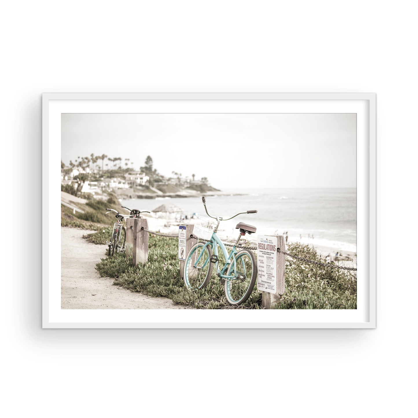 Fine art of horizontal landscape print by Southern California photographer Kim Dybczak of Salt Creek Prints, the image shows a light blue pastel beach cruiser leaning against a wooden post and chain fence. Another bicycle is visible behind it. The beach is in the background with people and a beach hut, located in Windansea Beach in La Jolla, California. Photograph in framed minimalist mock up