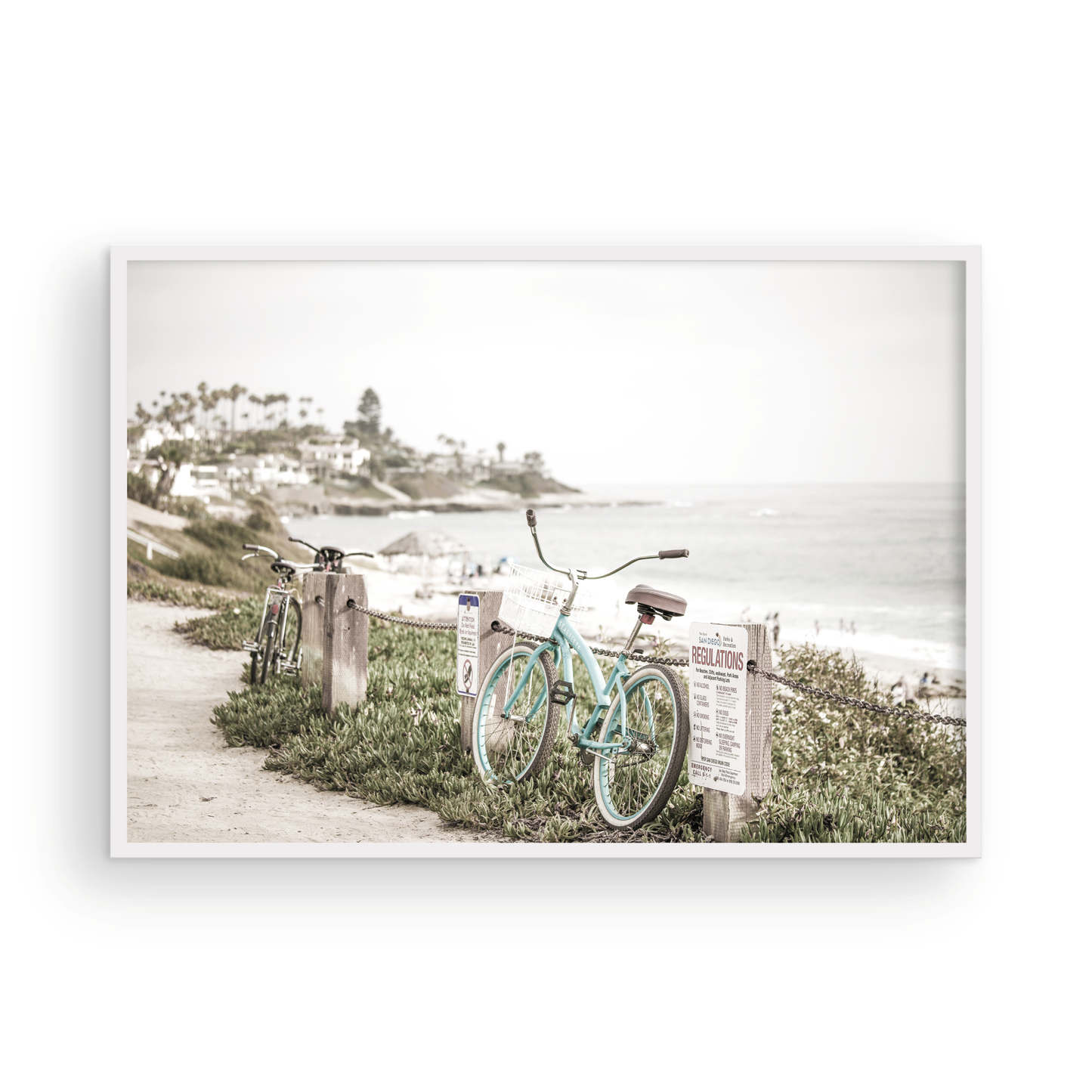 Fine art of horizontal landscape print by Southern California photographer Kim Dybczak of Salt Creek Prints, the image shows a light blue pastel beach cruiser leaning against a wooden post and chain fence. Another bicycle is visible behind it. The beach is in the background with people and a beach hut, located in Windansea Beach in La Jolla, California. Photograph in framed minimalist mock up
