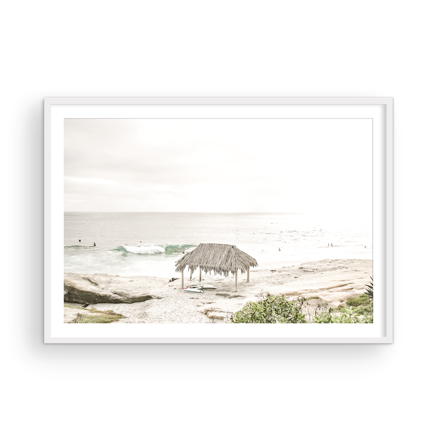 Fine art of WindanSea Beach, La Jolla California horizontal landscape print by Southern California photographer Kim Dybczak of Salt Creek Prints, featuring a bright, slightly desaturated image shows Windansea Beach in La Jolla, California. A thatched beach hut stands on the sandy shore in the left foreground. Further down the beach, a surfer carrying a teal surfboard walks towards the ocean where several other surfers are visible in the water. Photograph in minimalist framed mock up