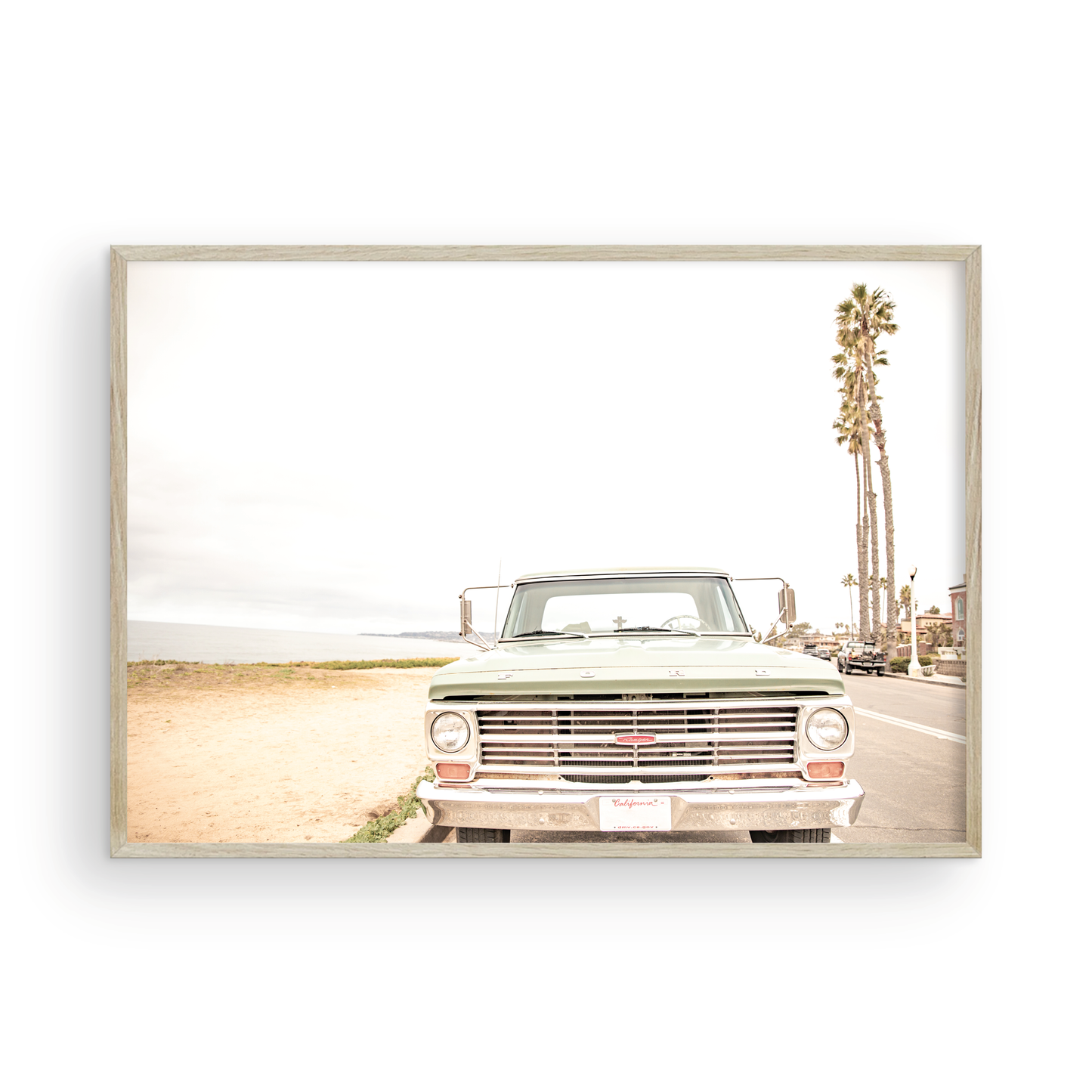 Nostalgic Southern California beach art featuring a classic light green Ford F-Series pickup truck parked by the San Diego coast. This horizontal surf print captures the vintage vehicle with a backdrop of iconic tall palm trees, a hazy beach, and the ocean, embodying surf vibes and perfect for coastal farmhouse decor. Photograph in minimalist framed mock up.