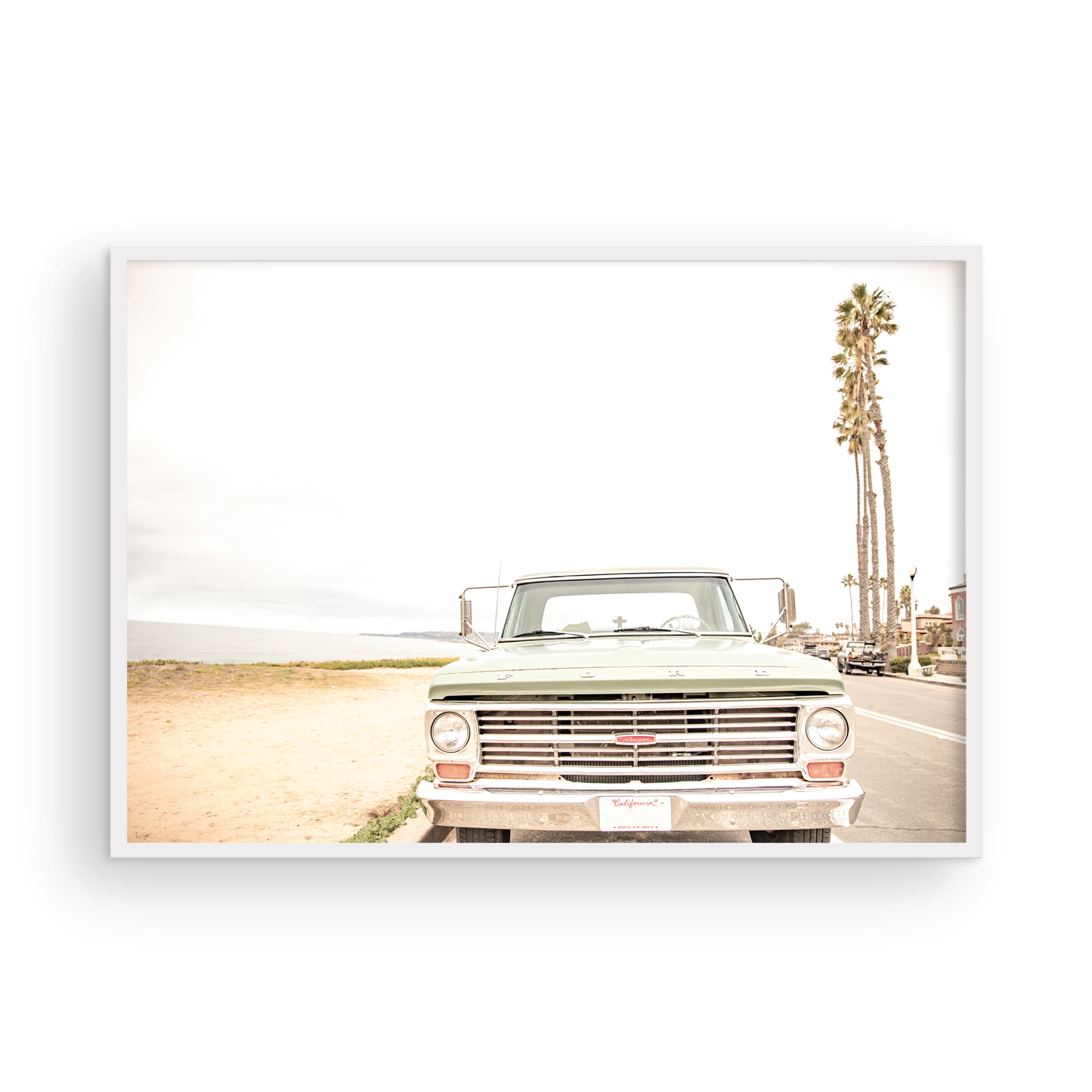 Nostalgic Southern California beach art featuring a classic light green Ford F-Series pickup truck parked by the San Diego coast. This horizontal surf print captures the vintage vehicle with a backdrop of iconic tall palm trees, a hazy beach, and the ocean, embodying surf vibes and perfect for coastal farmhouse decor. Photograph in minimalist framed mock up.