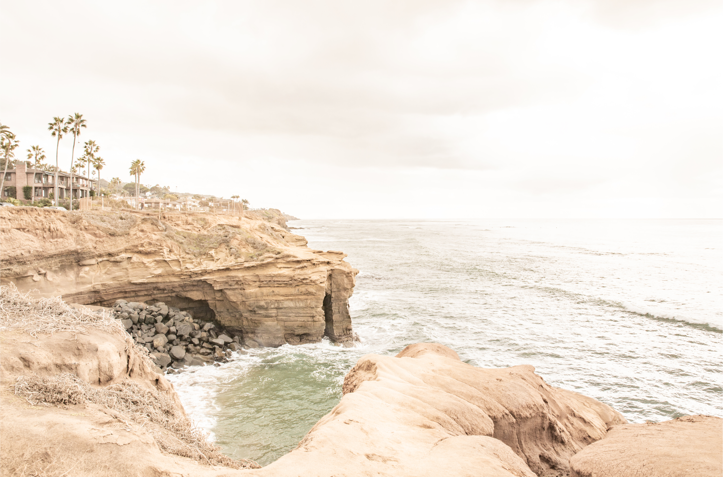Fine art of Sunset Cliffs, San Diego California horizontal print of hazy high-angle landscape beach photography captures a light blue ocean meeting tall tan, rocky, brown cliffs rise as iconic coastal scenery.