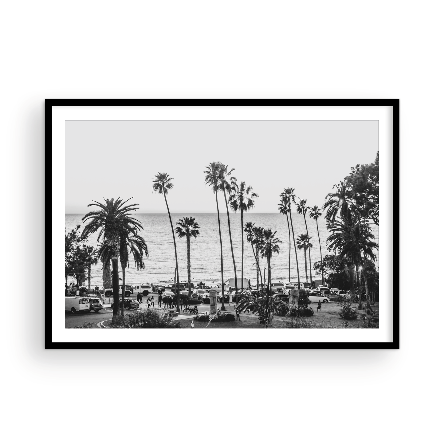 A classic black and white horizontal print of Swamis, the iconic San Diego surf spot, showcasing a panoramic view from above. This piece of Southern California travel photography features dozens of silhouetted palm trees against a bright sky, with a lively beach scene of cars and people below, stretching towards the ocean. Perfect as timeless beach art or neutral coastal decor for a surf vibe. Photograph in minimalist framed mock up.