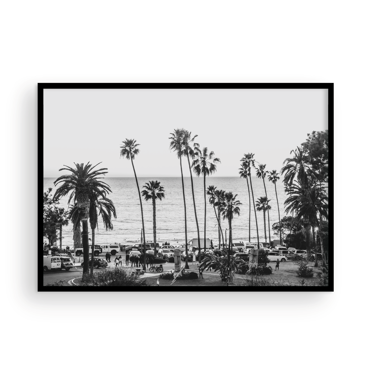 A classic black and white horizontal print of Swamis, the iconic San Diego surf spot, showcasing a panoramic view from above. This piece of Southern California travel photography features dozens of silhouetted palm trees against a bright sky, with a lively beach scene of cars and people below, stretching towards the ocean. Perfect as timeless beach art or neutral coastal decor for a surf vibe. Photograph in minimalist framed mock up.