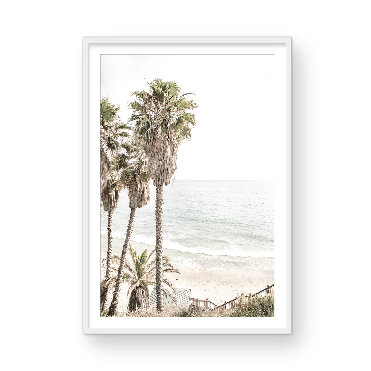 Dreamy coastal palm tree print featuring several tall palm trees with shaggy trunks, next to a staircase down to a tranquil beach. This vertical tropical print is captured at Swamis Beach on the Southern California coast, is bathed in bright, hazy sunlight, creating a pastel beach art effect. Evokes serene summer vibes, perfect for beach house decor. Photograph in minimalist surf art mock up.