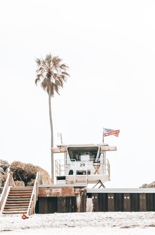 A photograph featuring an American flag on a lifeguard tower with a palm tree in the background, taken at a beach in Del Mar, San Diego.