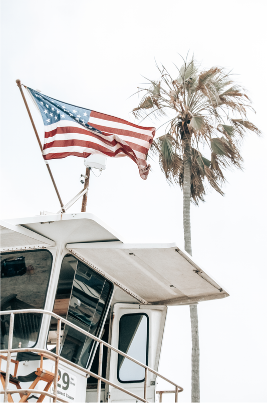 An American flag attached to a lifeguard tower with palm trees in the background, captured in California with a clear sky in the top half of the image.