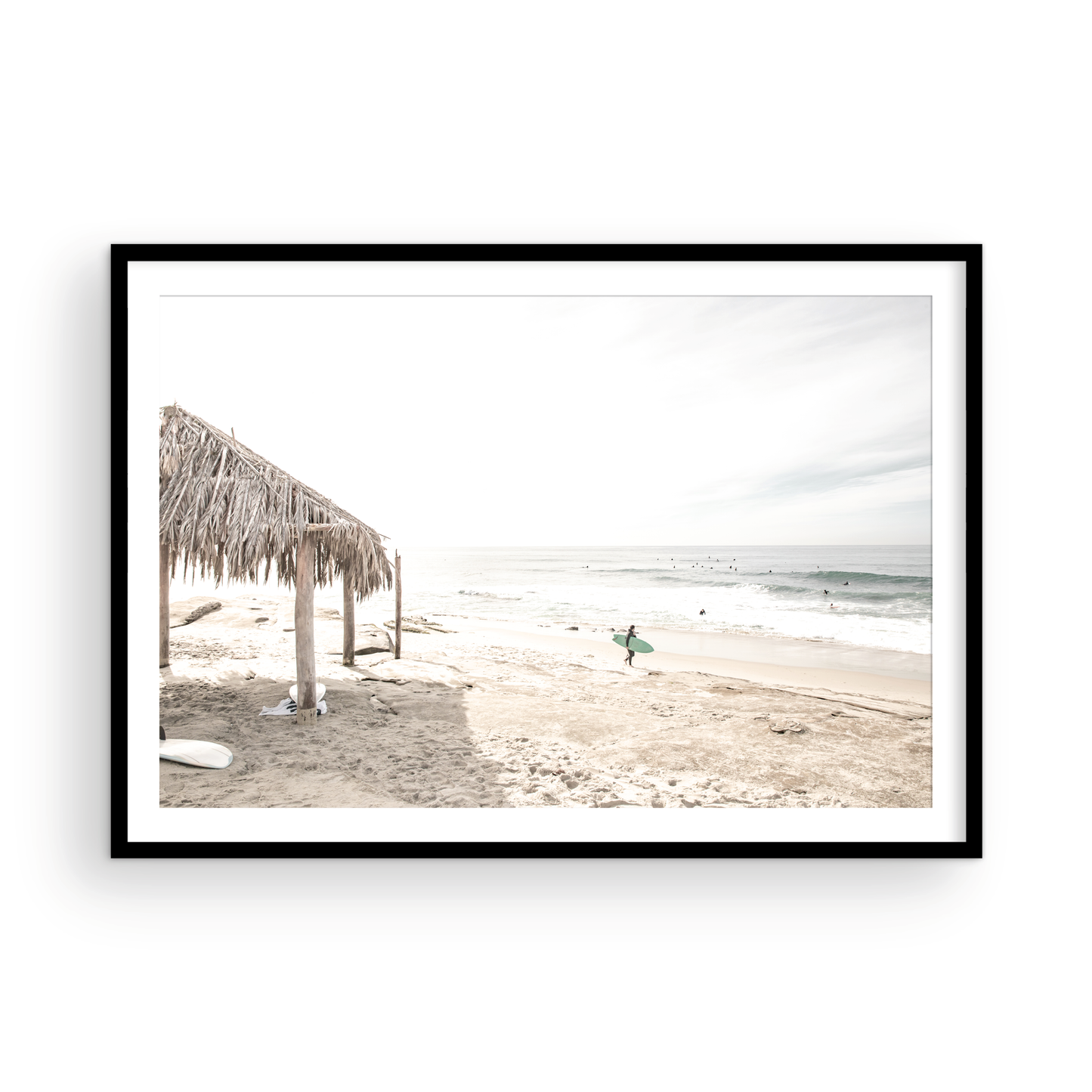Fine art of WindanSea Beach, La Jolla California horizontal landscape print by Southern California photographer Kim Dybczak of Salt Creek Prints, featuring a bright, slightly desaturated image shows Windansea Beach in La Jolla, California. A thatched beach hut stands on the sandy shore in the left foreground. Further down the beach, a surfer carrying a teal surfboard walks towards the ocean where several other surfers are visible in the water. Photograph in minimalist framed mock up