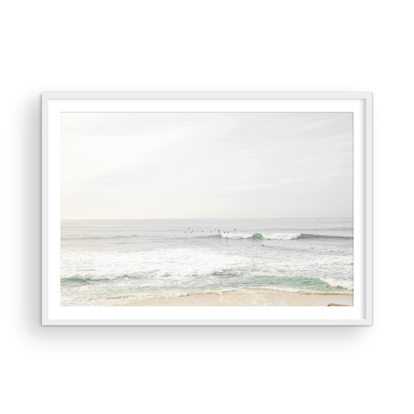 Fine art of horizontal landscape print by Southern California photographer Kim Dybczak of Salt Creek Prints, featuring a bright image captures a pale sandy beach giving way to a vast, light gray ocean with gentle waves. Several surfers are scattered across the water, some sitting on their boards and others riding small waves. The sky is a uniform, pale white, creating a soft, overcast atmosphere, located in Windansea Beach in La Jolla, California. Photograph in framed minimalist mock up