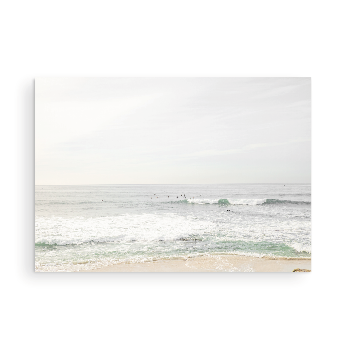 Fine art of horizontal landscape print by Southern California photographer Kim Dybczak of Salt Creek Prints, featuring a bright image captures a pale sandy beach giving way to a vast, light gray ocean with gentle waves. Several surfers are scattered across the water, some sitting on their boards and others riding small waves. The sky is a uniform, pale white, creating a soft, overcast atmosphere, located in Windansea Beach in La Jolla, California. Photograph in unframed minimalist mock up