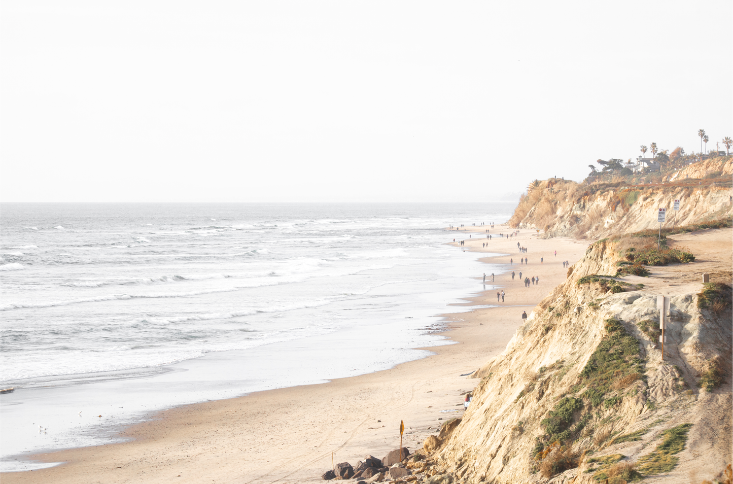 Fine art of Torrey Pines, San Diego California horizontal print of hazy high-angle landscape beach photography captures a light blue ocean meeting a sandy beach with many beach goers. To the right, tall tan and brown cliffs rise as iconic coastal scenery.