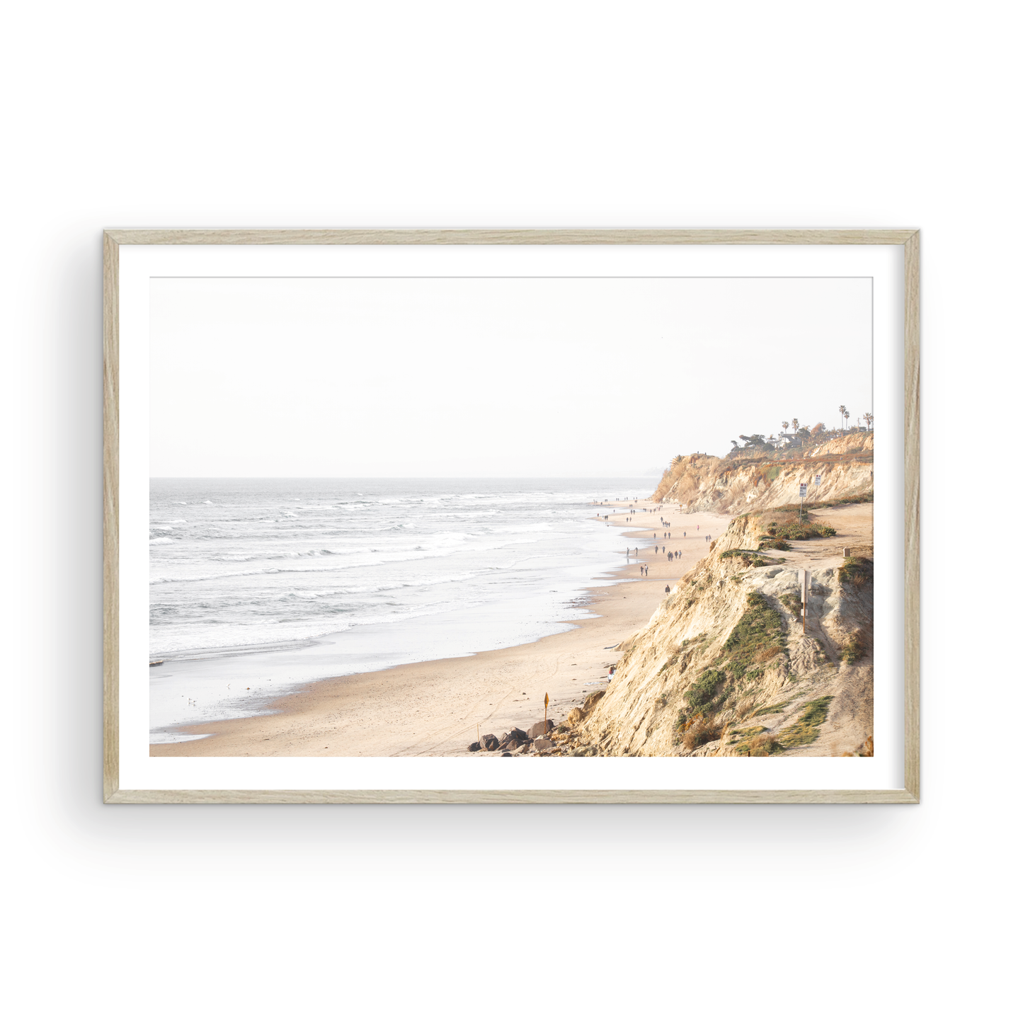 Fine art of Torrey Pines, San Diego California horizontal print of hazy high-angle landscape beach photography captures a light blue ocean meeting a sandy beach with many beach goers. To the right, tall tan and brown cliffs rise as iconic coastal scenery. Photograph in minimalist framed mock up.