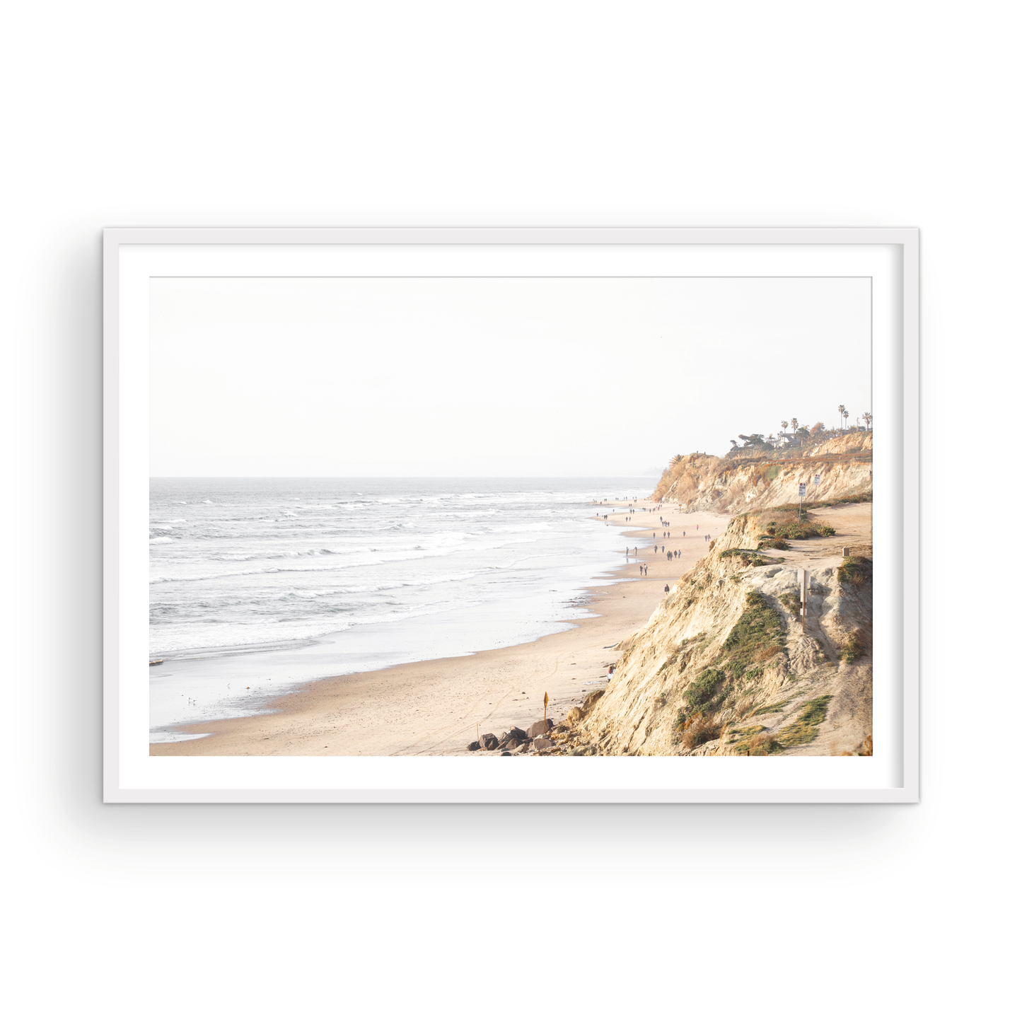 Fine art of Torrey Pines, San Diego California horizontal print of hazy high-angle landscape beach photography captures a light blue ocean meeting a sandy beach with many beach goers. To the right, tall tan and brown cliffs rise as iconic coastal scenery. Photograph in minimalist framed mock up.