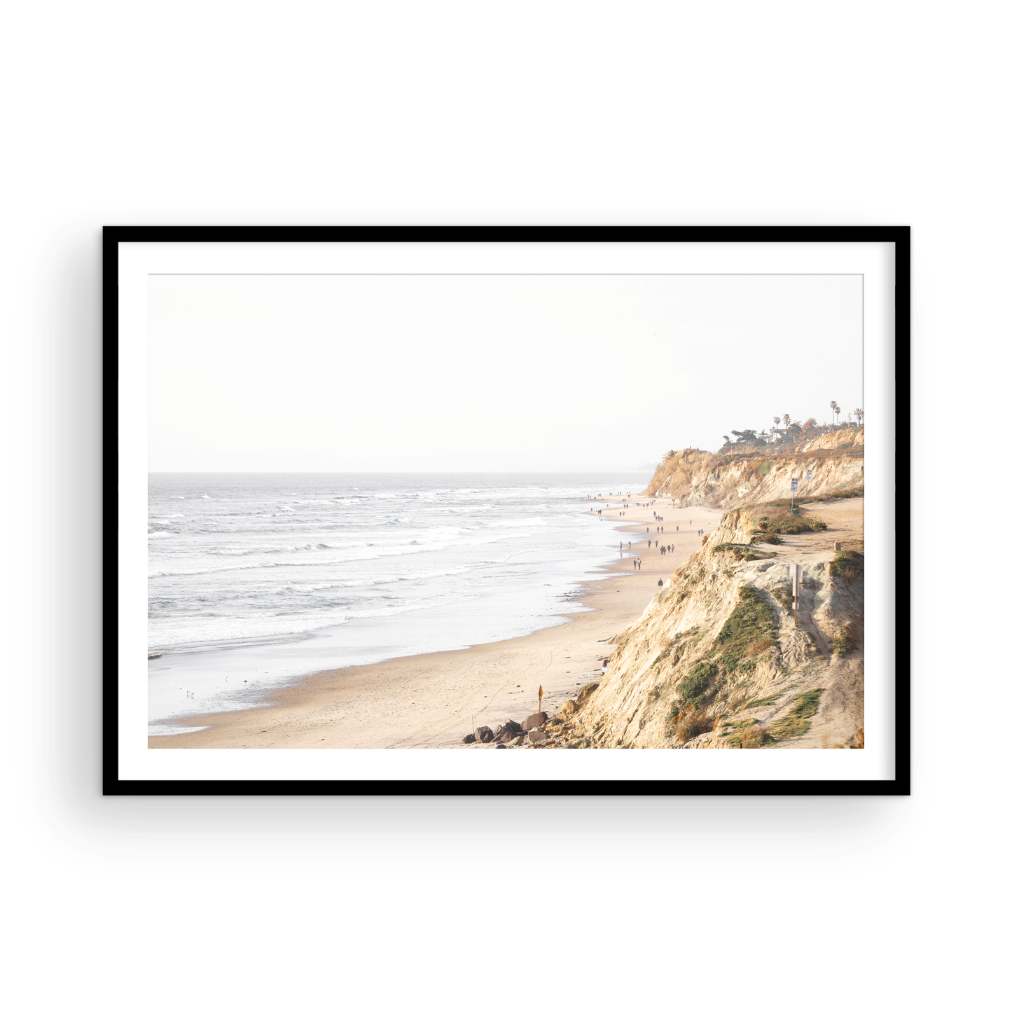 Fine art of Torrey Pines, San Diego California horizontal print of hazy high-angle landscape beach photography captures a light blue ocean meeting a sandy beach with many beach goers. To the right, tall tan and brown cliffs rise as iconic coastal scenery. Photograph in minimalist framed mock up.