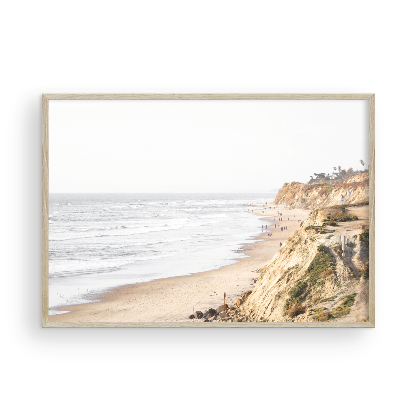 Fine art of Torrey Pines, San Diego California horizontal print of hazy high-angle landscape beach photography captures a light blue ocean meeting a sandy beach with many beach goers. To the right, tall tan and brown cliffs rise as iconic coastal scenery. Photograph in minimalist framed mock up.