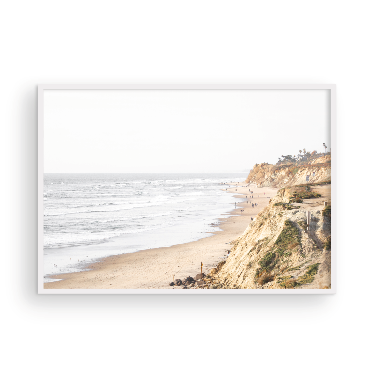 Fine art of Torrey Pines, San Diego California horizontal print of hazy high-angle landscape beach photography captures a light blue ocean meeting a sandy beach with many beach goers. To the right, tall tan and brown cliffs rise as iconic coastal scenery. Photograph in minimalist framed mock up.