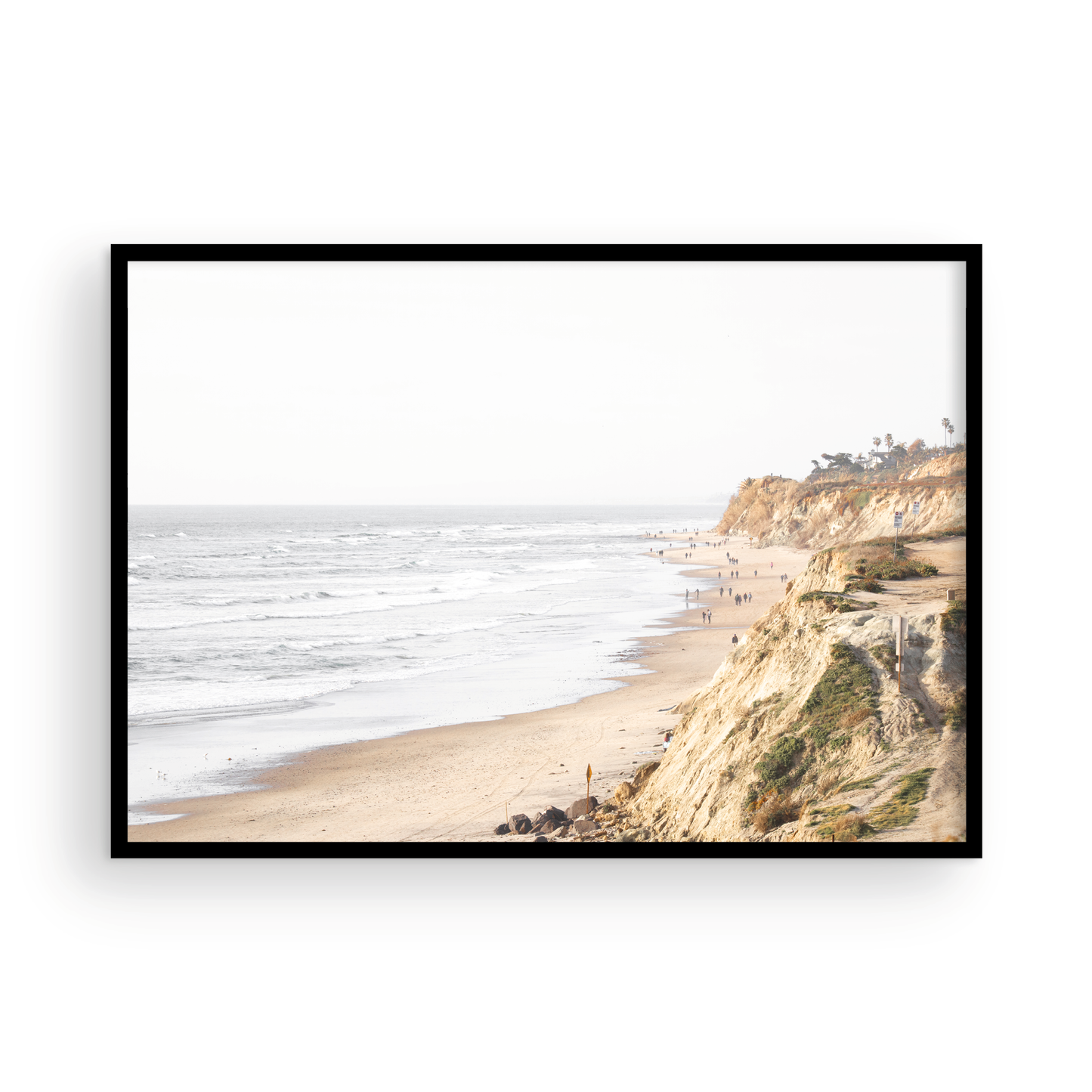 Fine art of Torrey Pines, San Diego California horizontal print of hazy high-angle landscape beach photography captures a light blue ocean meeting a sandy beach with many beach goers. To the right, tall tan and brown cliffs rise as iconic coastal scenery. Photograph in minimalist framed mock up.