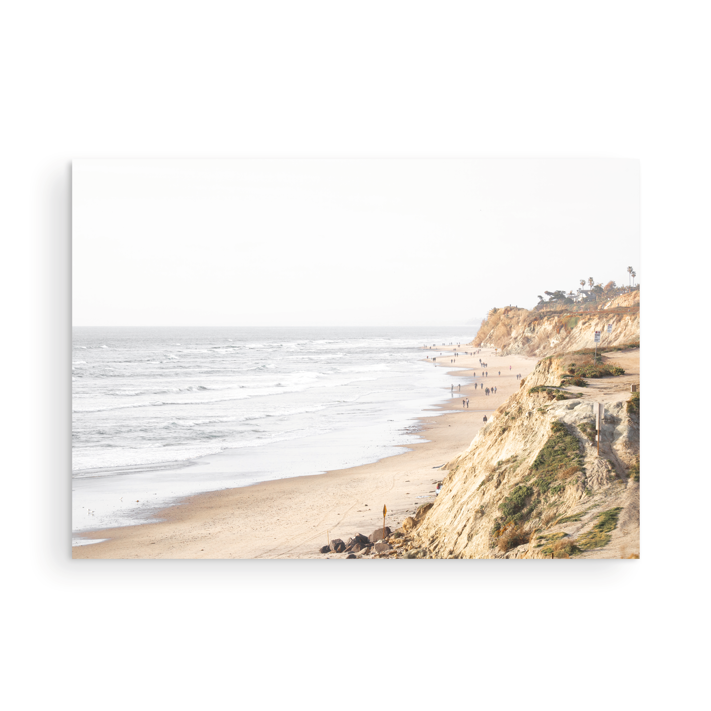 Fine art of Torrey Pines, San Diego California horizontal print of hazy high-angle landscape beach photography captures a light blue ocean meeting a sandy beach with many beach goers. To the right, tall tan and brown cliffs rise as iconic coastal scenery. Photograph in minimalist unframed mock up.