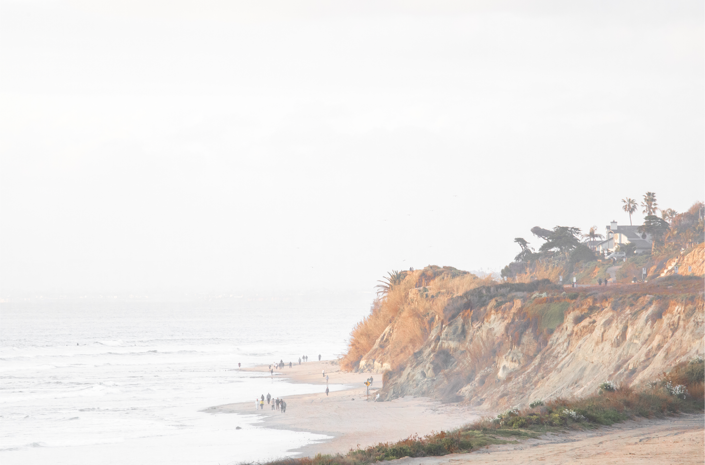 Fine art of Torrey Pines, San Diego California horizontal print of hazy high-angle landscape beach photography captures a light blue ocean meeting a sandy beach with many beach goers. To the right, tall tan and brown cliffs rise as iconic coastal scenery.