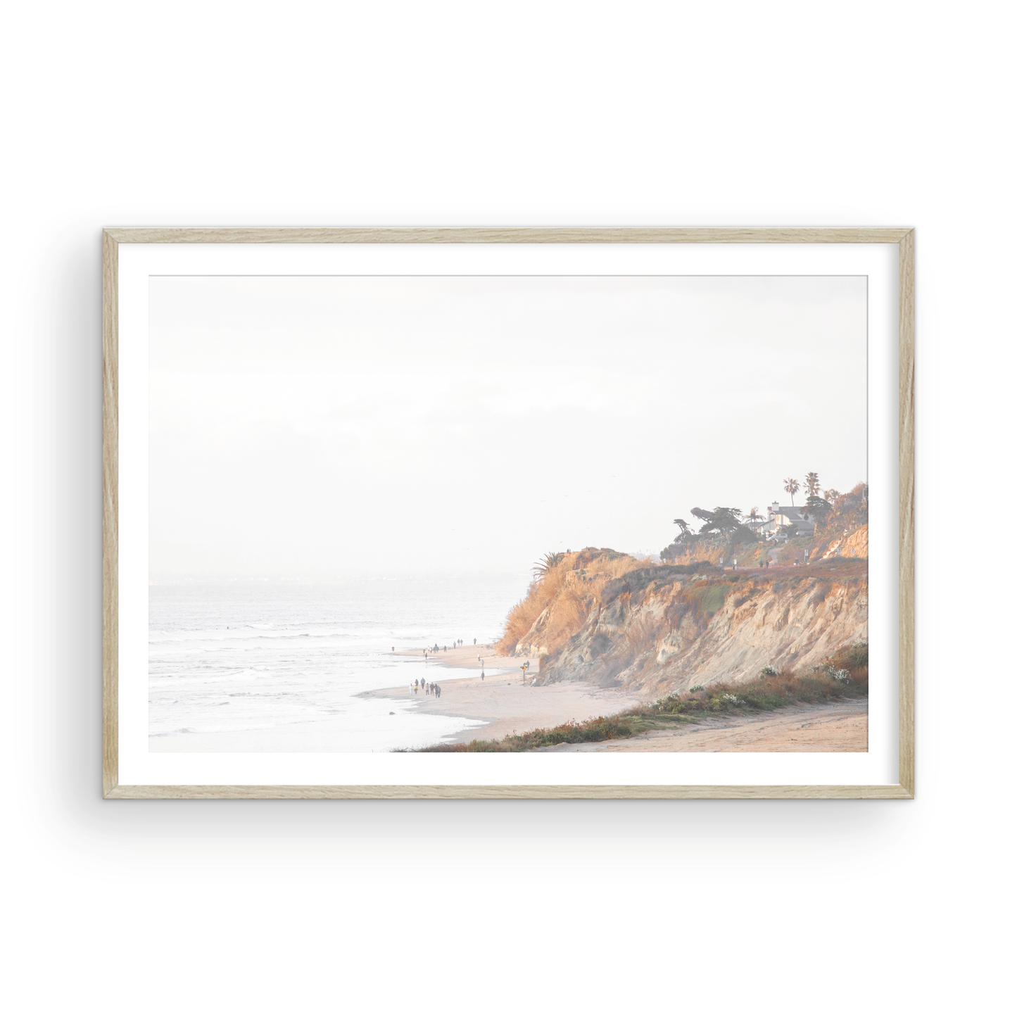 Fine art of Torrey Pines, San Diego California horizontal print of hazy high-angle landscape beach photography captures a light blue ocean meeting a sandy beach with many beach goers. To the right, tall tan and brown cliffs rise as iconic coastal scenery. Photograph in minimalist framed mock up.