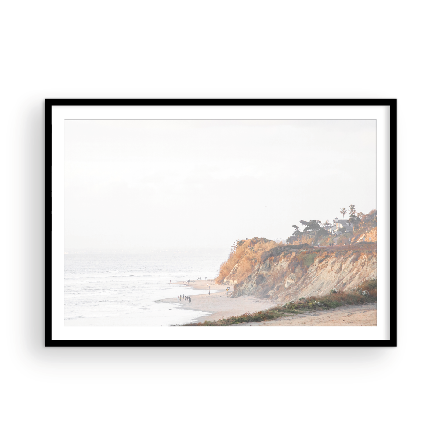 Fine art of Torrey Pines, San Diego California horizontal print of hazy high-angle landscape beach photography captures a light blue ocean meeting a sandy beach with many beach goers. To the right, tall tan and brown cliffs rise as iconic coastal scenery. Photograph in minimalist framed mock up.