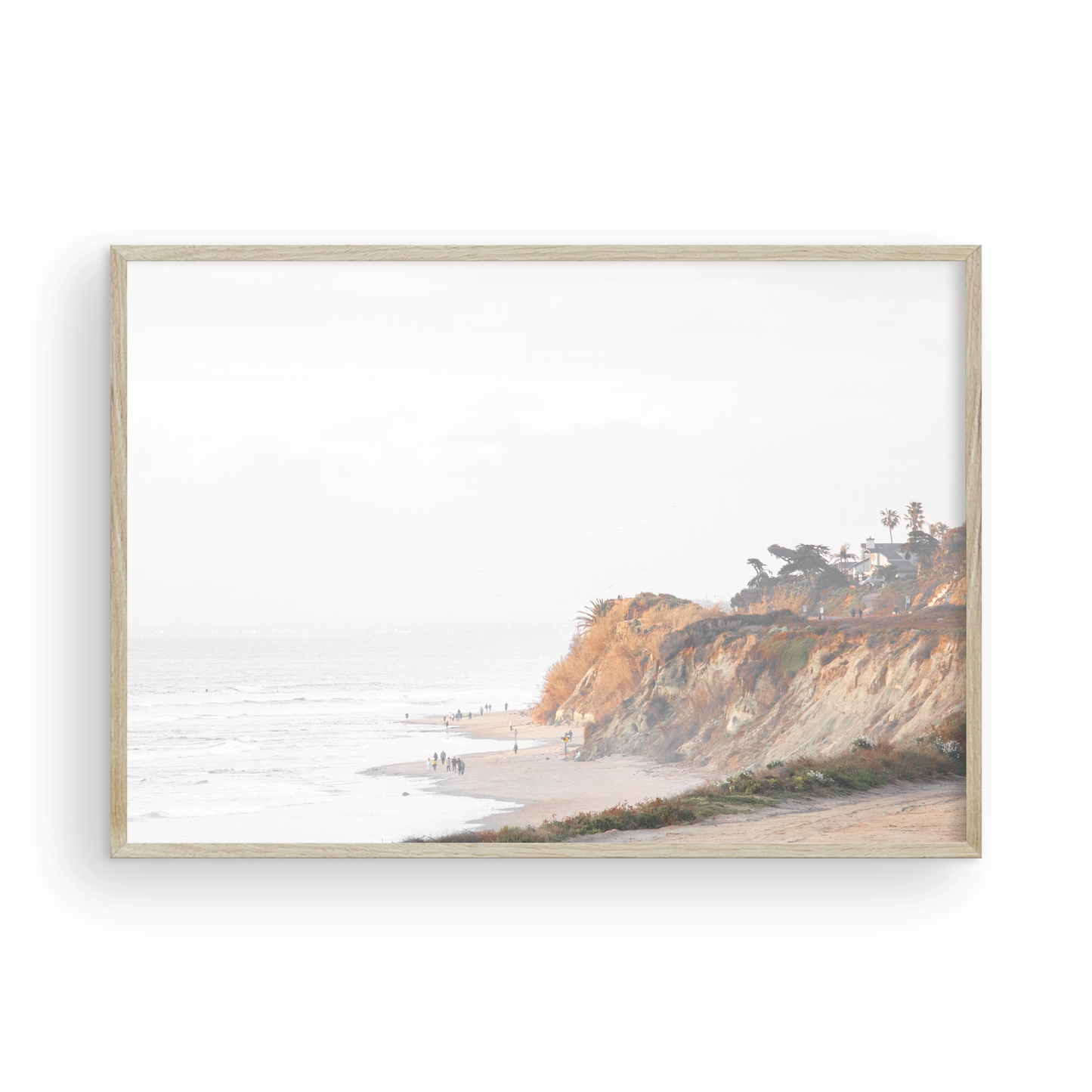 Fine art of Torrey Pines, San Diego California horizontal print of hazy high-angle landscape beach photography captures a light blue ocean meeting a sandy beach with many beach goers. To the right, tall tan and brown cliffs rise as iconic coastal scenery. Photograph in minimalist framed mock up.