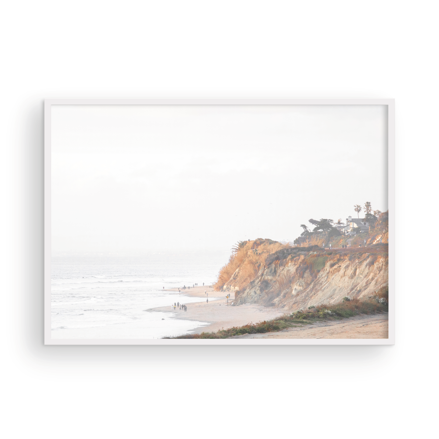 Fine art of Torrey Pines, San Diego California horizontal print of hazy high-angle landscape beach photography captures a light blue ocean meeting a sandy beach with many beach goers. To the right, tall tan and brown cliffs rise as iconic coastal scenery. Photograph in minimalist framed mock up.