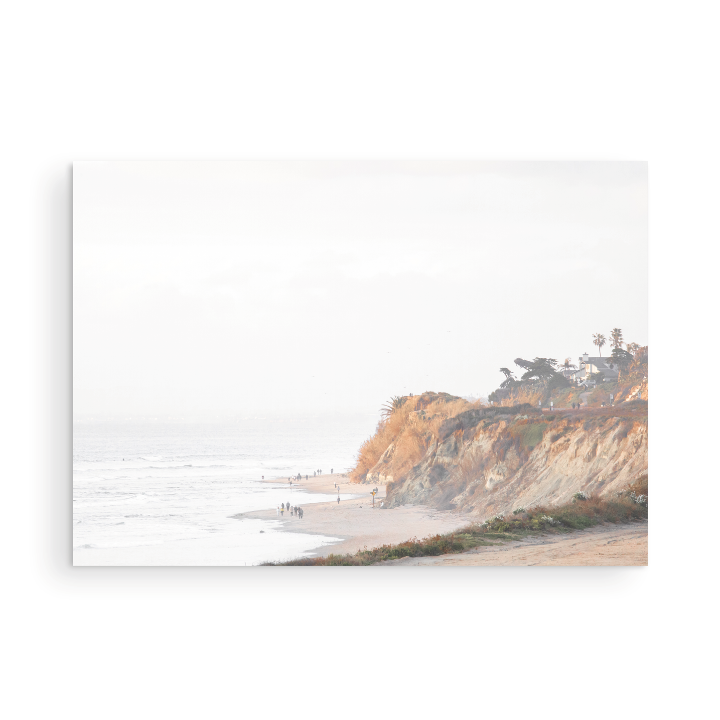 Fine art of Torrey Pines, San Diego California horizontal print of hazy high-angle landscape beach photography captures a light blue ocean meeting a sandy beach with many beach goers. To the right, tall tan and brown cliffs rise as iconic coastal scenery. Photograph in minimalist unframed mock up.