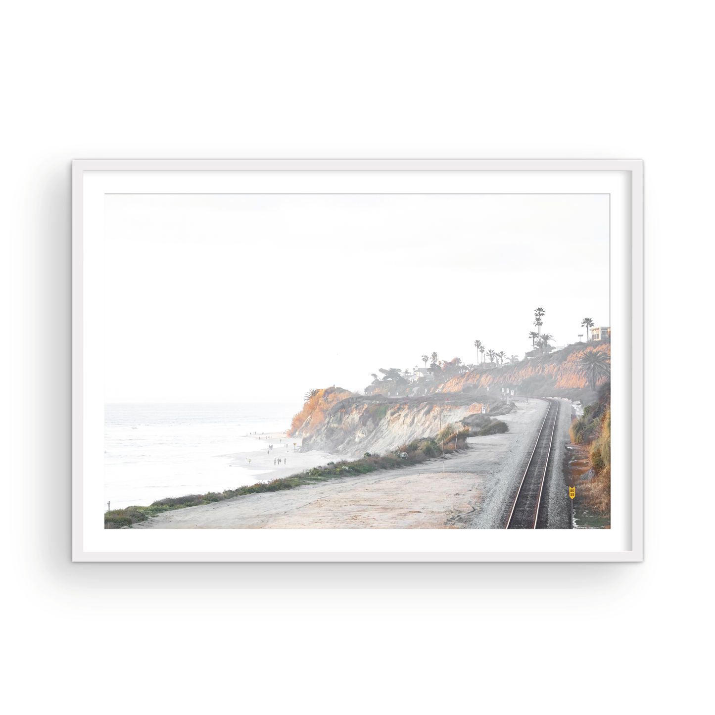 A slightly hazy, wide Landscape Coastal Photography shot shows a set of train tracks curving along a coastal cliffside, San Diego Coast. To the left, a sandy beach meets a light blue ocean waves with gentle waves, where a few small figures of people are visible. The cliffs are varied in color, with sections of tan, white, and hints of orange, topped with greenery and a few Palm Trees. The sky is a muted white, perfect for Minimalist Surf Wall Art. Photograph in minimalist mock up