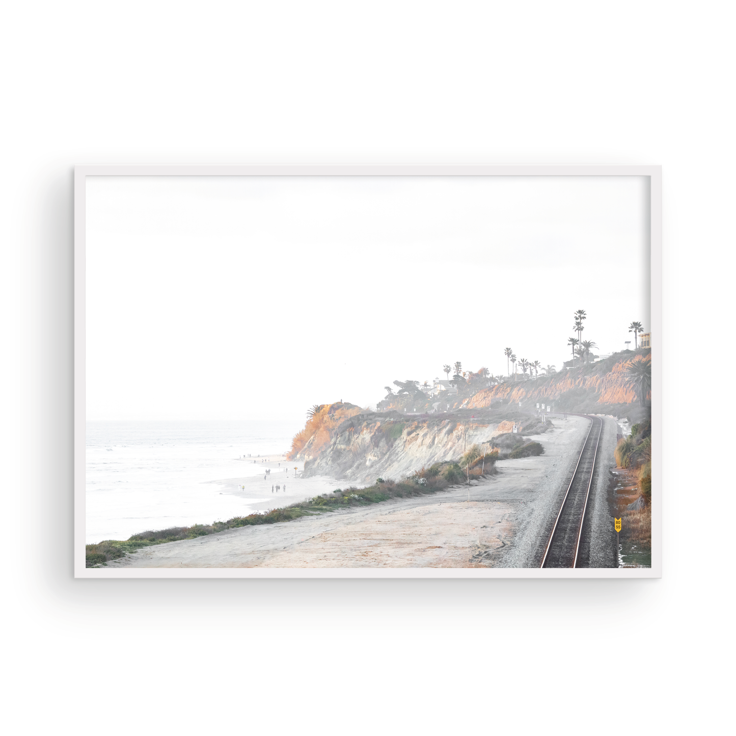 A slightly hazy, wide Landscape Coastal Photography shot shows a set of train tracks curving along a coastal cliffside, San Diego Coast. To the left, a sandy beach meets a light blue ocean waves with gentle waves, where a few small figures of people are visible. The cliffs are varied in color, with sections of tan, white, and hints of orange, topped with greenery and a few Palm Trees. The sky is a muted white, perfect for Minimalist Surf Wall Art. Photograph in minimalist mock up